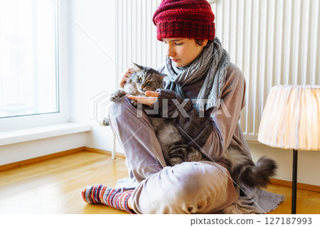 woman in knitted hat, scarf, mittens, hugging cat, sits at home near radiator 127187993