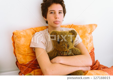 Portrait teenage girl, in bed with her favorite toy, grimaces, shows tongue 127188015