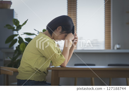 一位 30 多歲的女性在家裡的餐桌旁抱著頭,拿著智慧型手機,側面 一位 30 多歲的女性在家裡的餐桌旁抱著頭,拿著智慧型手機,側面 127188744