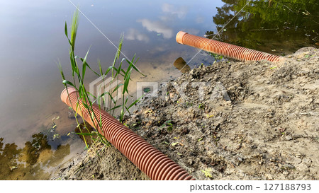 Ribbed drainage pipes descend into a tranquil pond from a worn, sandy shoreline with minimal plant life. Pollution or wastewater outflow . 127188793