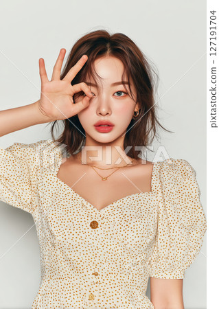 Young woman with short brown hair, wearing polka dot dress, making OK hand gesture over her eye, looking confident 127191704