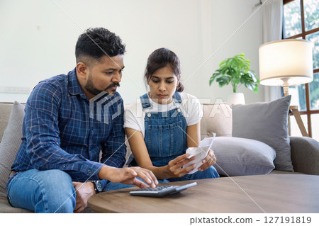 Indian married couple calculating bills, discussing planning budget together, serious wife and husband looking at calculator, checking finances Indian married couple calculating bills, discussing planning budget together, serious wife and husband looking at calculator, checking finances 127191819