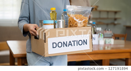 charity, donation and volunteering concept. Volunteer prepare foodstuff box containing food to donate to people poor, disaster victim at home charity, donation and volunteering concept. Volunteer prepare foodstuff box containing food to donate to people poor, disaster victim at home 127191894