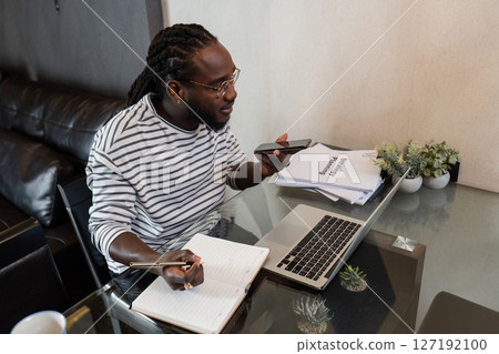 Young African American business man working with laptop and document on desk and talking on mobile at home Young African American business man working with laptop and document on desk and talking on mobile at home 127192100