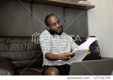African American business man working with document and laptop while talking on mobile at home on couch. work from home concept 127192109