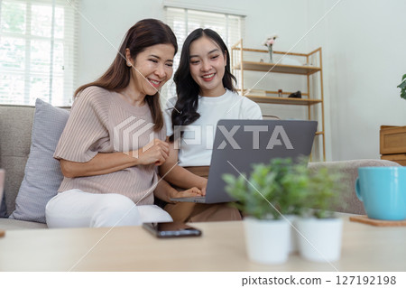 Mother and adult daughter sitting on the sofa together, mother and daughter using laptop to surf website together 127192198