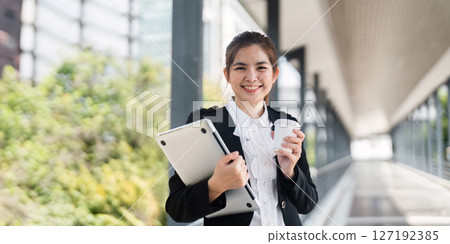 Young busy successful Asian business woman, professional businesswoman holding laptop and holding coffee cup standing or walking on urban business center 127192385
