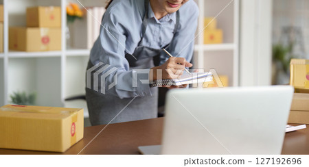 Small Business Operations. Entrepreneur managing orders at a desk with laptop and packages. Small Business Operations. Entrepreneur managing orders at a desk with laptop and packages. 127192696