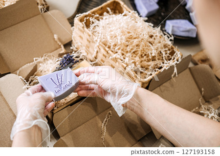 Gloved hands hold lavender soap above shredded filler and open kraft packaging. Product presentation, boutique branding, handmade business, artisan soap design Gloved hands hold lavender soap above shredded filler and open kraft packaging. Product presentation, boutique branding, handmade business, artisan soap design 127193158