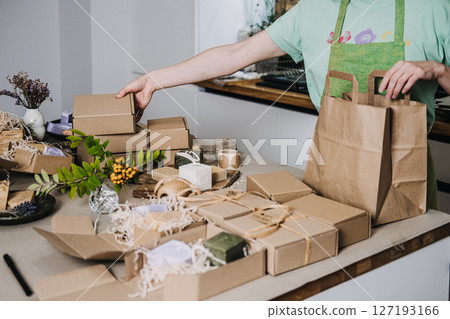 Person places kraft box into a paper bag at a workspace with handmade soap packaging. Sustainable packaging, biodegradable wrap, eco unboxing, zero waste branding 127193166