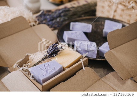 Lavender soaps are arranged in open kraft boxes with shredded filler on a workspace table. Plastic-free box, shredded kraft filler, sustainable wrap, eco-friendly presentation Lavender soaps are arranged in open kraft boxes with shredded filler on a workspace table. Plastic-free box, shredded kraft filler, sustainable wrap, eco-friendly presentation 127193169