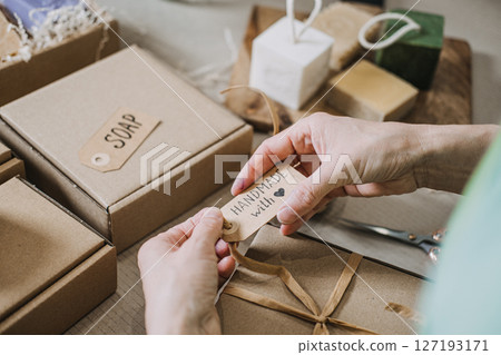 Hands attach a handmade soap tag to a kraft gift box beside artisanal skincare bars. Handmade with care, ethical entrepreneurship, slow made, purpose-driven business 127193171
