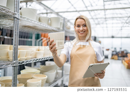A cheerful woman in an apron holds up a flowerpot in a plant shop, with shelves of pots in the background. A cheerful woman in an apron holds up a flowerpot in a plant shop, with shelves of pots in the background. 127193531