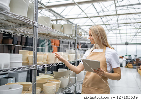 A woman in an apron looks at flowerpots in a shop. She is holding a tablet. 127193539
