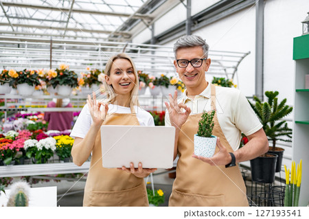 Two Caucasian florists show a plant and laptop, giving an OK sign, in their vibrant shop. The employees look happy and satisfied with their work. 127193541