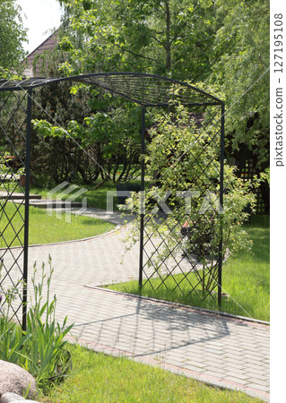 Wrought iron arch, support for flowers in park, backyard. Nature and plants in summer. Concrete tile walkway. Wrought iron arch, support for flowers in park, backyard. Nature and plants in summer. Concrete tile walkway. 127195108