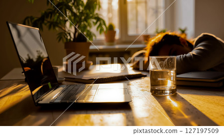A person sleeping on a desk next to a laptop computer and a glass of water 127197509