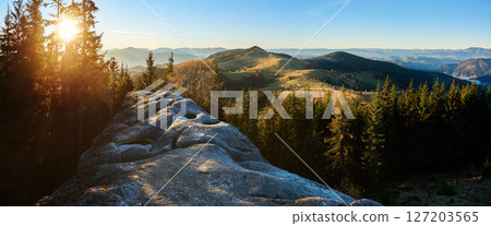 Panoramic image of sunrise over Pysanyi Kamin in Carpathians, Ukraine. Sun's rays pierce through tall pine trees, illuminating rocky foreground with water-filled depressions. 127203565