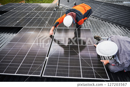 Professional technicians adjusting solar PV panels. Solar system installation on roof of house. Top view of two male workers in halmets and work overalls securing solar battery. Professional technicians adjusting solar PV panels. Solar system installation on roof of house. Top view of two male workers in halmets and work overalls securing solar battery. 127203567