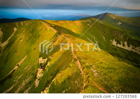 Breathtaking aerial view of green, rugged mountain peaks under dramatic sky. Scene captures contrast between vibrant landscape and moody clouds, with sunlight breaking through. Carpathians, Chornogora 127203568
