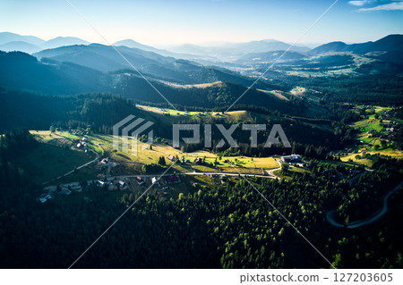Aerial view of densely forested mountains stretching into distance under clear blue sky with scattered clouds. Landscape reveals valleys and distant fields, showcasing natural beauty of region. Aerial view of densely forested mountains stretching into distance under clear blue sky with scattered clouds. Landscape reveals valleys and distant fields, showcasing natural beauty of region. 127203605