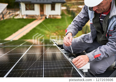 Man worker mounting photovoltaic solar panels on roof of house. Close up view of engineer installing solar module system with help of hex key. Concept of alternative, renewable energy. Man worker mounting photovoltaic solar panels on roof of house. Close up view of engineer installing solar module system with help of hex key. Concept of alternative, renewable energy. 127203606