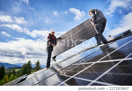 Roofers building solar panel system on roof of house. Men workers in helmets carrying photovoltaic solar module outdoors. Concept of alternative and renewable energy. Roofers building solar panel system on roof of house. Men workers in helmets carrying photovoltaic solar module outdoors. Concept of alternative and renewable energy. 127203607