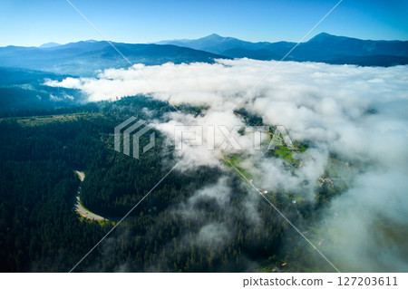 Mesmerizing aerial view of landscape blanketed in fluffy white clouds. Lush green valleys and winding roads peek through, while distant blue mountains stand majestically under bright, clear sky. Mesmerizing aerial view of landscape blanketed in fluffy white clouds. Lush green valleys and winding roads peek through, while distant blue mountains stand majestically under bright, clear sky. 127203611