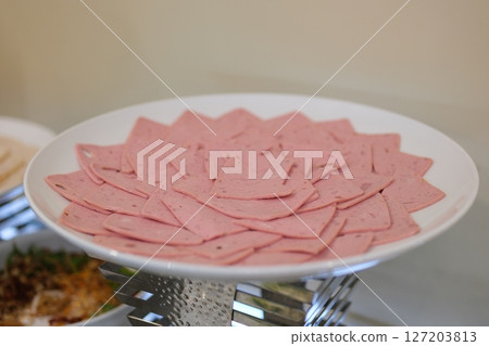 A plate of sliced, pink meat product rests on a red surface, showcasing its smooth texture and distinctive shape. A plate of sliced, pink meat product rests on a red surface, showcasing its smooth texture and distinctive shape. 127203813