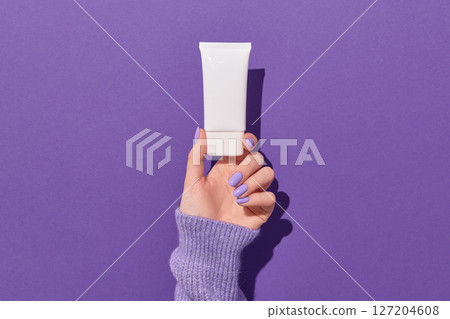 Womans hand holding white tube on violet background. Skin health care concept. Womans hand holding white tube on violet background. Skin health care concept. 127204608