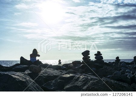 A girl sits on the coastal stones on the ocean shore in the rays of a bright setting sun A girl sits on the coastal stones on the ocean shore in the rays of a bright setting sun 127206141