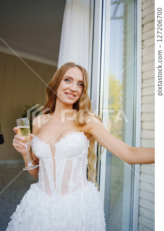 A bride with long blonde hair with a glass of champagne by an open window. A bride with long blonde hair with a glass of champagne by an open window. 127206700