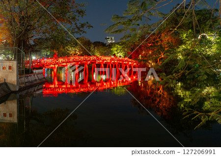 A vivid night scene captures the iconic The Huc Bridge in Hanoi, Vietnam, reflecting in the tranquil waters. The red bridge glows under the lights. A vivid night scene captures the iconic The Huc Bridge in Hanoi, Vietnam, reflecting in the tranquil waters. The red bridge glows under the lights. 127206991