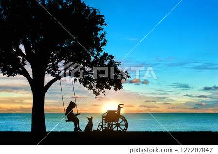 Girl schoolgirl invalid sitting on a swing under a tree by the sea, reading a book, beside a wheelchair and her dog 127207047