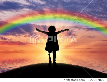 A happy child is a disabled girl with a prosthetic leg in the open air on the background of sea sunset and rainbow 127207072
