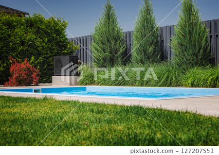 Modern heat pump installed next to a backyard pool, used for heating water, surrounded by green lawn, conifers, and privacy fence in a landscaped garden. 127207551