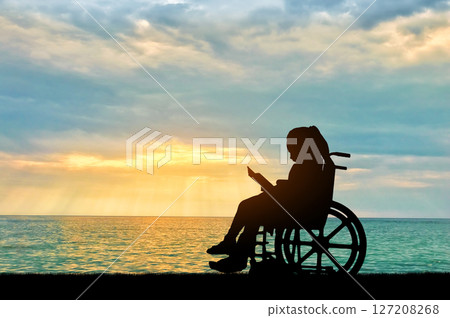 Silhouette of a disabled child girl sitting in a wheelchair reading a book on a sea sunset background Silhouette of a disabled child girl sitting in a wheelchair reading a book on a sea sunset background 127208268