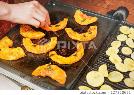 Bright orange butternut squash pieces grilled on electric grill, blurred woman hand moving pieces of vegetables over hot plate 127210651