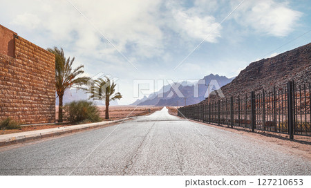 Long straight road with desert hills in distance, brick wall and iron fence on sides. Scenery at the entrance to Wadi Rum protected area of Jordan 127210653