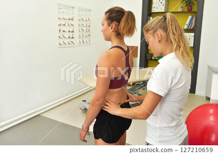 Young female physiotherapist exercising with her patient, examining lower back muscles from behind 127210659