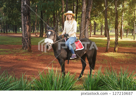 Young woman in shirt and straw hat, riding dark brown horse in the park, blurred tall pine trees in background 127210687