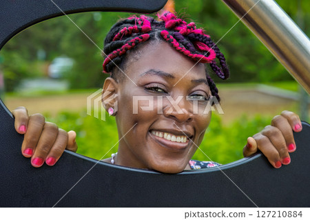 Joyful Black female kindergarten educator with colorful braids at children's playground, perfect for childcare and early education. 127210884