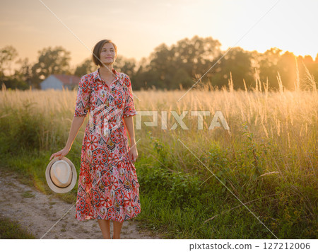 Young woman walking through picturesque European field in late summer. Golden sunlight, lush greenery, and serene rural atmosphere create peaceful countryside scene. Young woman walking through picturesque European field in late summer. Golden sunlight, lush greenery, and serene rural atmosphere create peaceful countryside scene. 127212006