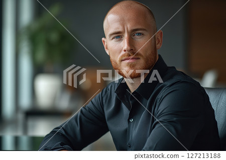 Young attractive male executive in black shirt with bald head, light stubble and blue eyes sitting at table in leather chair in modern office. Generated by artificial intelligence 127213188