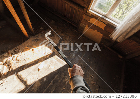 Person holding a crowbar in an old, derelict wooden room with sunlight 127213390