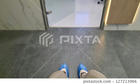 Waiting at a clinic entrance. First-person view of feet in blue shoe covers. Healthcare, medical visit concept. Clean and sterile environment. 127213966