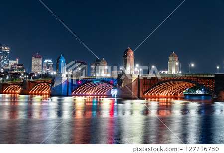 Night view of the Longfellow Bridge in Boston glowing with orange lighting, arching over the Charles River with reflections shimmering on the water and the city skyline in the background Night view of the Longfellow Bridge in Boston glowing with orange lighting, arching over the Charles River with reflections shimmering on the water and the city skyline in the background 127216730