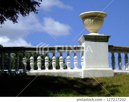 A white railing features smooth balusters and supports a decorative urn. This charming detail is part of an idyllic park landscape under a clear blue sky, creating a peaceful atmosphere. 127217202