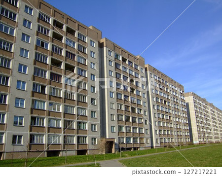 Tall residential buildings stretch across the landscape under a bright blue sky. The well-maintained structure features numerous windows that reflect sunlight, creating a vivid urban ambiance. 127217221