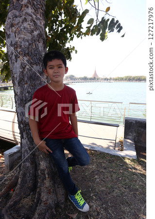 Young boy in a red shirt standing next to a tree by a tranquil lakeside, showcasing nature and childhood in an outdoor setting with peaceful water and greenery Young boy in a red shirt standing next to a tree by a tranquil lakeside, showcasing nature and childhood in an outdoor setting with peaceful water and greenery 127217976